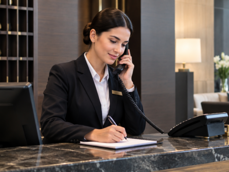 răspuns telefon recepție hotel într-o scenă cu recepționer la desk, vorbind calm la telefon și notând corect informațiile oaspetelui.